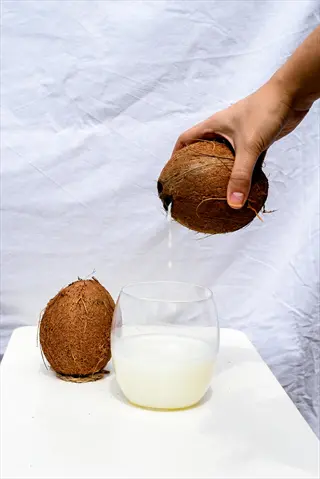 hand pouring coconut water from a fresh coconut into a glass (coconut water glass), with another whole coconut on a white table against a plain background