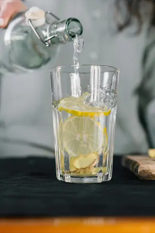 hand pouring water from glass bottle into a glass filled with lemon and ginger slices on a dark table surface