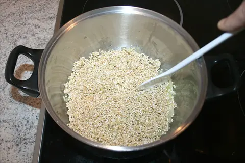 hand stirring cooked pearl barley in stainless steel pot on black stovetop with white spoon and kitchen countertop