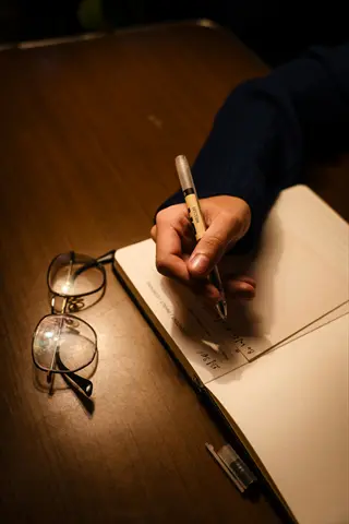 hand writing in open journal with pen, glasses resting nearby - visual of gratitude journaling practice