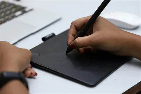 hands using stylus on graphics tablet for digital painting in a workspace with laptop and mouse, embodying digital art