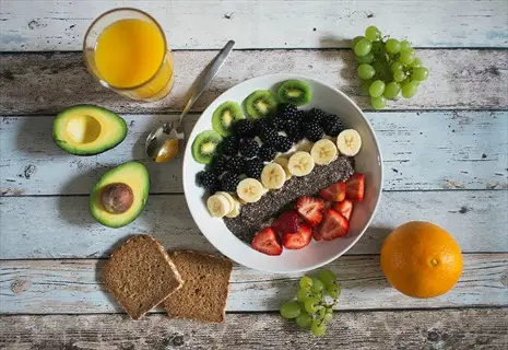 heavy metal detox foods flat lay: chia seed bowl layered with kiwi, blackberries, bananas, and strawberries; avocado halves, orange, green grapes, whole grain toast, and fresh orange juice on rustic wood