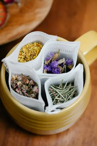 herbal tea assortment mugs: a yellow mug holds four distinct tea bags with dried botanicals (purple flowers, golden buds, brown petals, green herbs) for herbal tea hydration