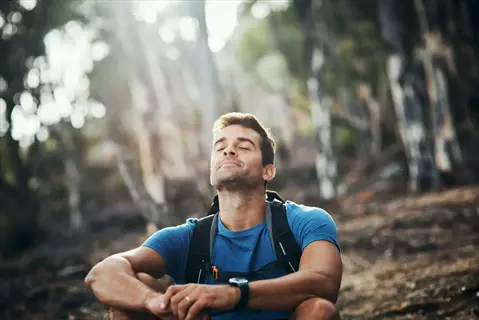 hiker practicing deep breathing outdoor in sunlit forest, eyes closed with peaceful expression amidst trees