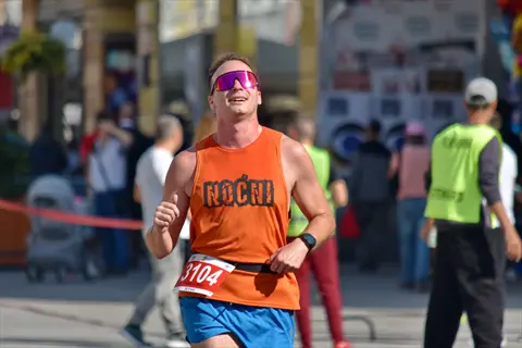 male athlete running endurance race in bright sunlight: wearing orange 'rocni' tank top, blue shorts, race bib 3104, sunglasses, and fitness watch, surrounded by other runners and spectators in urban setting