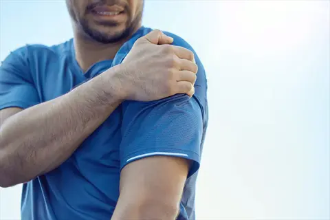 man holding shoulder to relieve muscle tension, demonstrating shoulder tension relief techniques against a clear sky background
