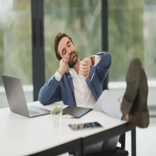 man in blue blazer relaxing at office desk with feet up, talking on phone while checking wristwatch