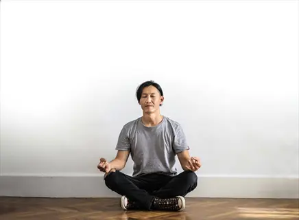 man meditating in cross-legged floor sitting position on wooden floor against white wall