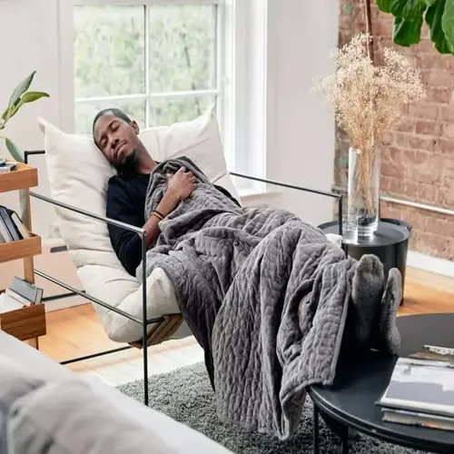 man resting under weighted blanket bed on modern chaise lounge in sunlit living room with brick walls and plants