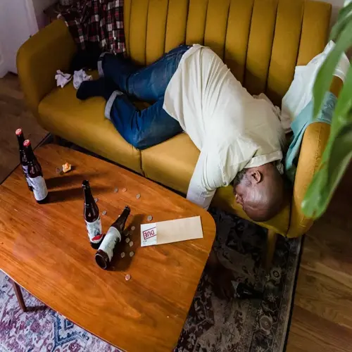 man sleeping on a couch after drinking, with beer bottles and scattered coins on the table, and a 'due' envelope visible
