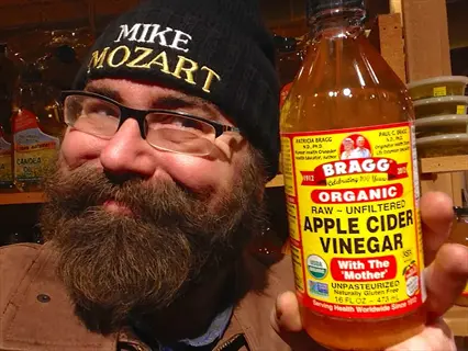 man with beard and glasses wearing 'mike mozart' beanie holds bragg organic apple cider vinegar bottle labeled 'with the mother' in store