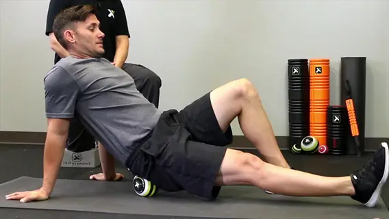 massage ball glutes: man using a green massage ball on his glutes and hips while seated on a yoga mat in a fitness studio
