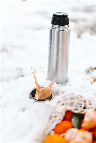 metallic insulated water bottle in snow with beverage splash and mesh fruit bag