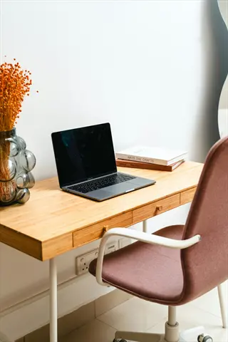 minimalist desk setup with wooden table, open laptop, stacked books, dried botanical arrangement in glass vase, and pink ergonomic chair in bright workspace