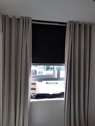 modern bedroom window with blackout curtains closed (light-blocking beige drapes and dark roller shade) for light management, revealing partial city view
