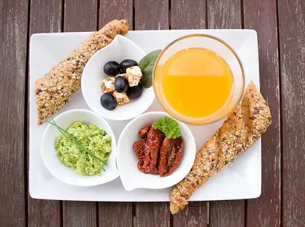 morning vitamins breakfast platter with whole grain bread, fresh orange juice, olives, feta cheese, avocado, and sun-dried tomatoes in white dishes on wooden table