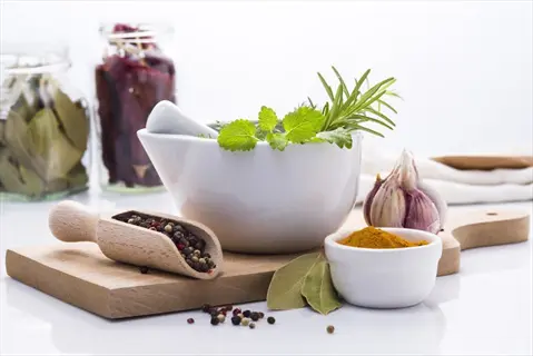 mortar with fresh rosemary, mint, and herbs surrounded by spices: peppercorns, turmeric powder, garlic, bay leaves, and jars of dried ingredients