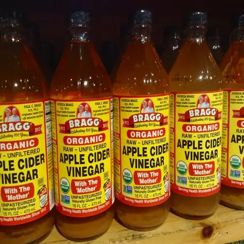 multiple bragg organic apple cider vinegar bottles with prominent labels showing text and founder portraits, displayed on a wooden retail shelf