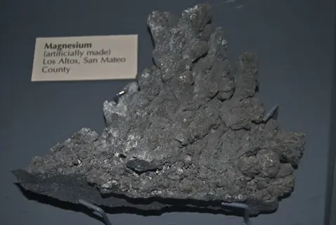 museum-displayed artificial magnesium mineral specimen with dendritic crystalline structure, shown with descriptive plaque reading 'magnesium (artificially made) los altos, san mateo county' on gray background