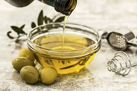 olive oil pour from bottle into glass bowl with fresh green olives and rosemary sprigs on rustic surface