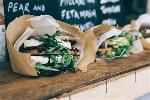 organic food safety at a market stall: wrapped sandwiches with fresh greens on a wooden table, chalkboard menu in background