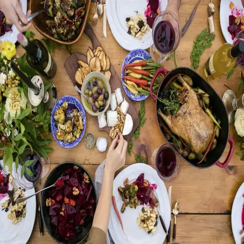 overhead view of a rustic wooden table spread with roasted poultry, beet salad, olives, cheese, and wine; a person eating slowly and mindfully reaches for nuts