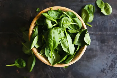 overhead view of raw spinach leaves overflowing a wooden bowl on a dark textured surface, with scattered leaves around