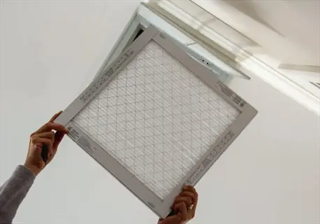person checking air quality by holding and inspecting an hvac air filter with visible mesh grid and printed text, against a light-colored ceiling background