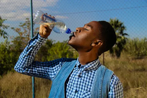 person drinking water from a 'bonaqua' bottle outdoors, wearing a blue checkered shirt and denim vest under sunny skies with trees in the background