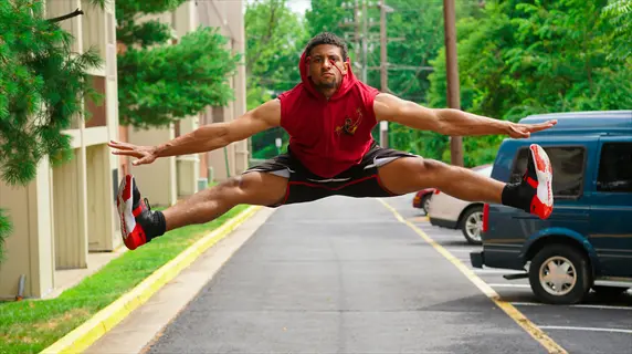person exercising outdoors: man performing mid-air split jump in suburban street setting with buildings, trees, and parked cars, demonstrating dynamic exercise synergy