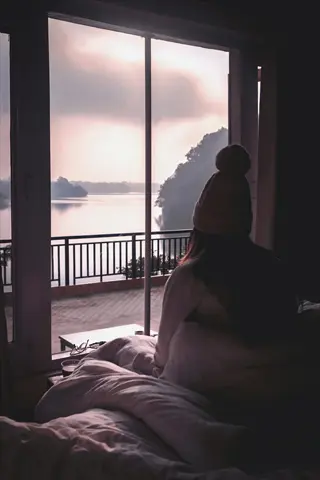 person in pom-pom hat sitting on bed in sunset-lit bedroom, gazing at serene lake view through large windows for circadian rhythm alignment