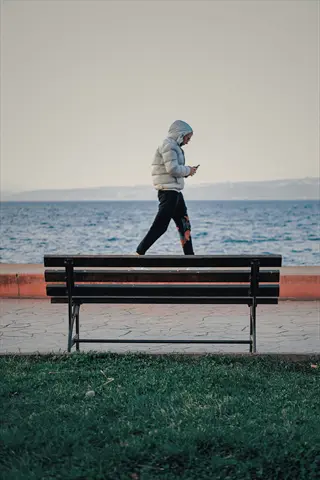 person in puffy hooded jacket stepping near a park bench by the sea while looking at a phone