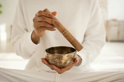 person in white clothing conducting singing bowl therapy with brass bowl and wooden mallet in bright serene room