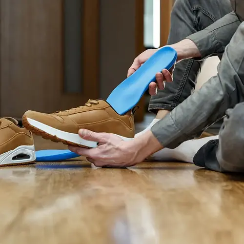 person inserting blue orthopedic insole into brown sneaker for comfortable foot arch support, on wooden floor with second shoe visible