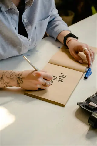 person journaling art: tattooed hand writing in 'art journal' notebook with pen on desk, art supplies visible, embodying self-reflection