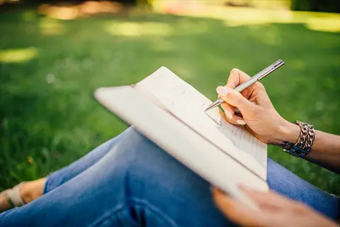 person journaling with pen in notebook on sunny grassy field, showcasing reflective writing practice