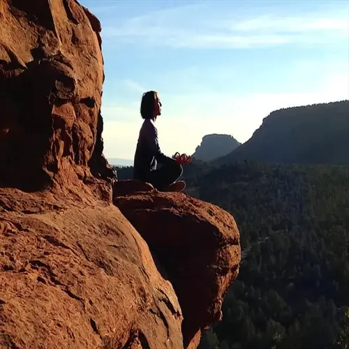 person meditating calmly on red rock cliff edge overlooking forested mountains at sunset