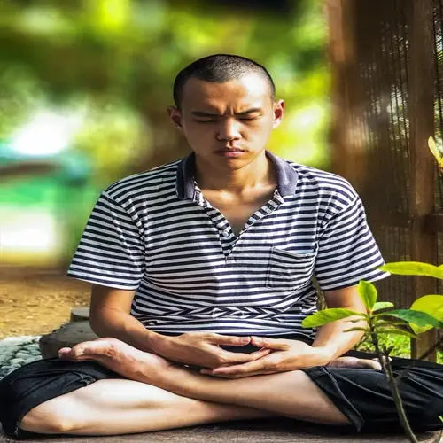 person meditating concentration in lotus position outdoors, eyes closed with hands on knees, wearing striped shirt in serene green blurred background