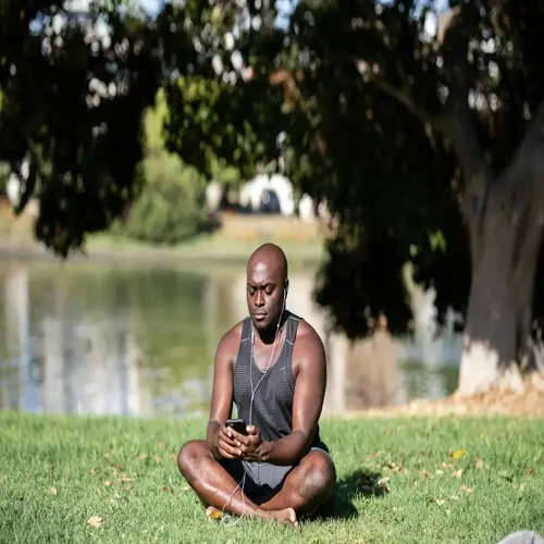 person meditating with smartphone in park by lake, cross-legged on grass under shady trees