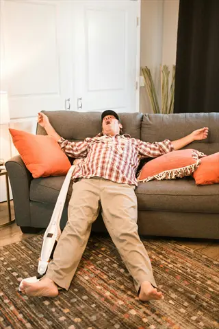 person napping on sofa with arms outstretched, yawn visible, surrounded by orange pillows and household items in a living room for napping strategies