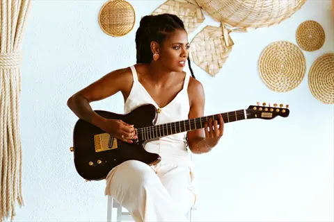 person playing guitar: woman in white dress with dreadlocks plays brown fender guitar against light blue wall with woven baskets decor