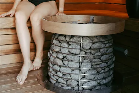 person relaxing near an electric sauna heater with stacked stone core and wooden frame in a sauna interior