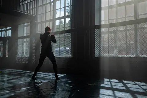 person shadow boxing in a dimly lit gym with dramatic sunlight streaming through industrial windows and chain-link fencing