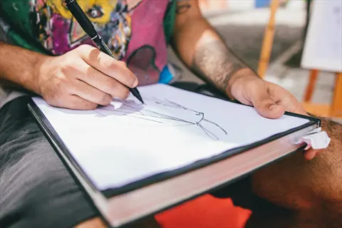 person sketching paper: tattooed hand drawing abstract line art with black pen in sketchbook, demonstrating drawing as a creative process