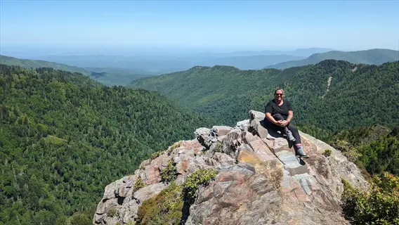 person smiling at mountain hiking trail summit with panoramic green valleys and distant peaks under clear blue sky