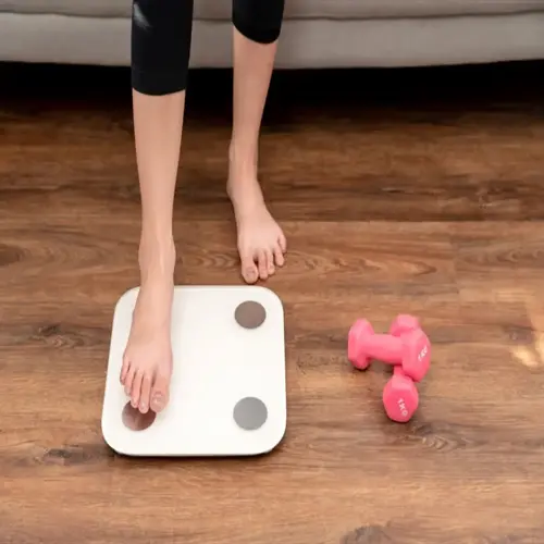 person stepping onto a modern body composition scale with pink dumbbells on wooden floor in living room