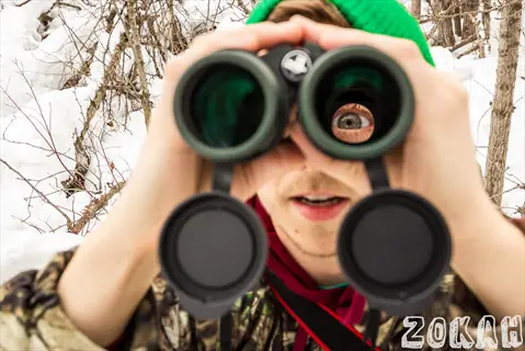 person using bird watching binoculars in snowy woodland with eye visible through lens and 'zokah' text watermark