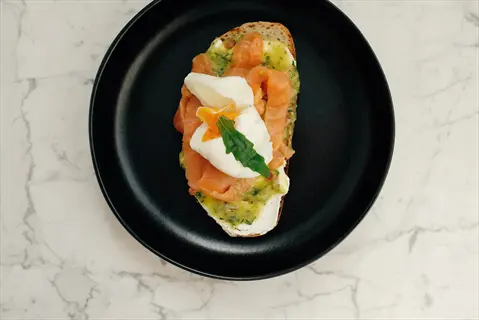 poached eggs with smoked salmon (fish) on an open-faced sandwich featuring green herb spread, served on a black plate against marble