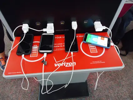 public verizon phone charging station with multiple smartphones connected to charging cables and power blocks in a busy environment