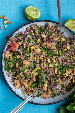 quinoa salad bowl: overhead view of a vibrant quinoa salad bowl with pistachios, fresh herbs, red onions, and lime wedges on a blue background